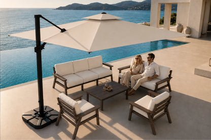 Outdoor patio furniture set by a poolside location with a couple sitting on a sofa under a large beige cantilever umbrella. The set includes a three-seat sofa, two armchairs, a square coffee table, and a black cantilever umbrella on a light stone deck with a blue ocean and mountains in the background.