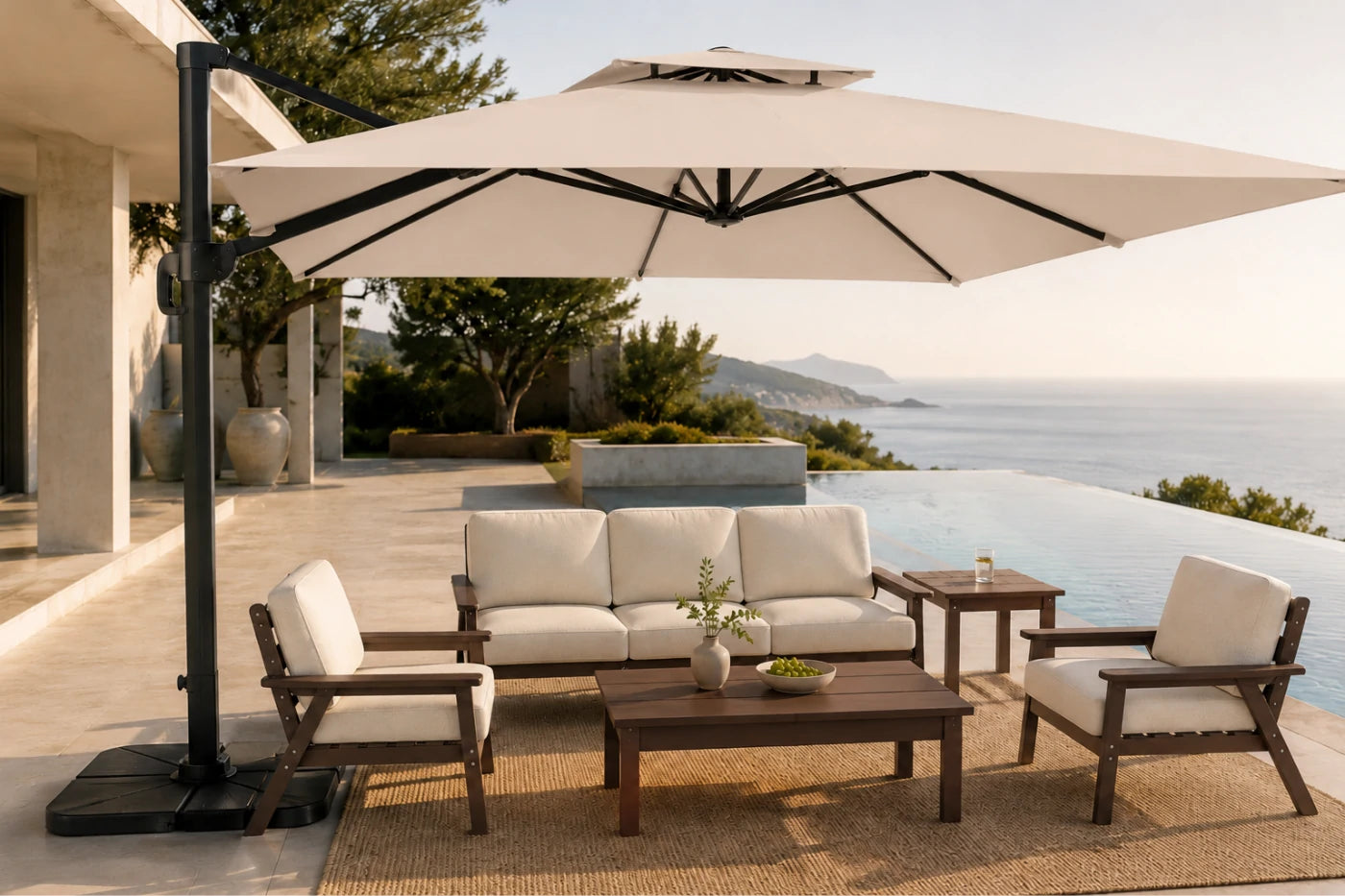 Outdoor patio furniture set with a beige patio umbrella, a three-seat sofa, two armchairs, and a coffee table on a woven rug beside an infinity pool overlooking the sea.