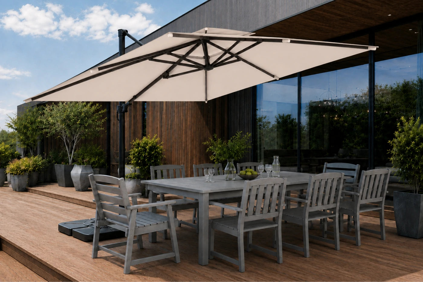 A large beige patio umbrella with a black frame is shown on a wooden deck next to a gray outdoor dining table and chairs, with a modern house and plants in the background.