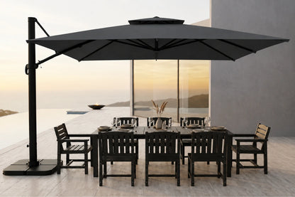 A large dark gray cantilever patio umbrella with a rectangular dining table and six matching black chairs, set on a light stone terrace overlooking an infinity pool and sunset seascape.