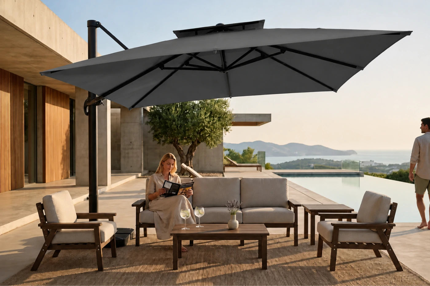 A large dark gray cantilever patio umbrella with a double canopy shade is set up on a modern outdoor terrace next to a swimming pool. The umbrella provides shade over a dark wood outdoor sofa, two matching armchairs with light beige cushions, and a low wooden coffee table.