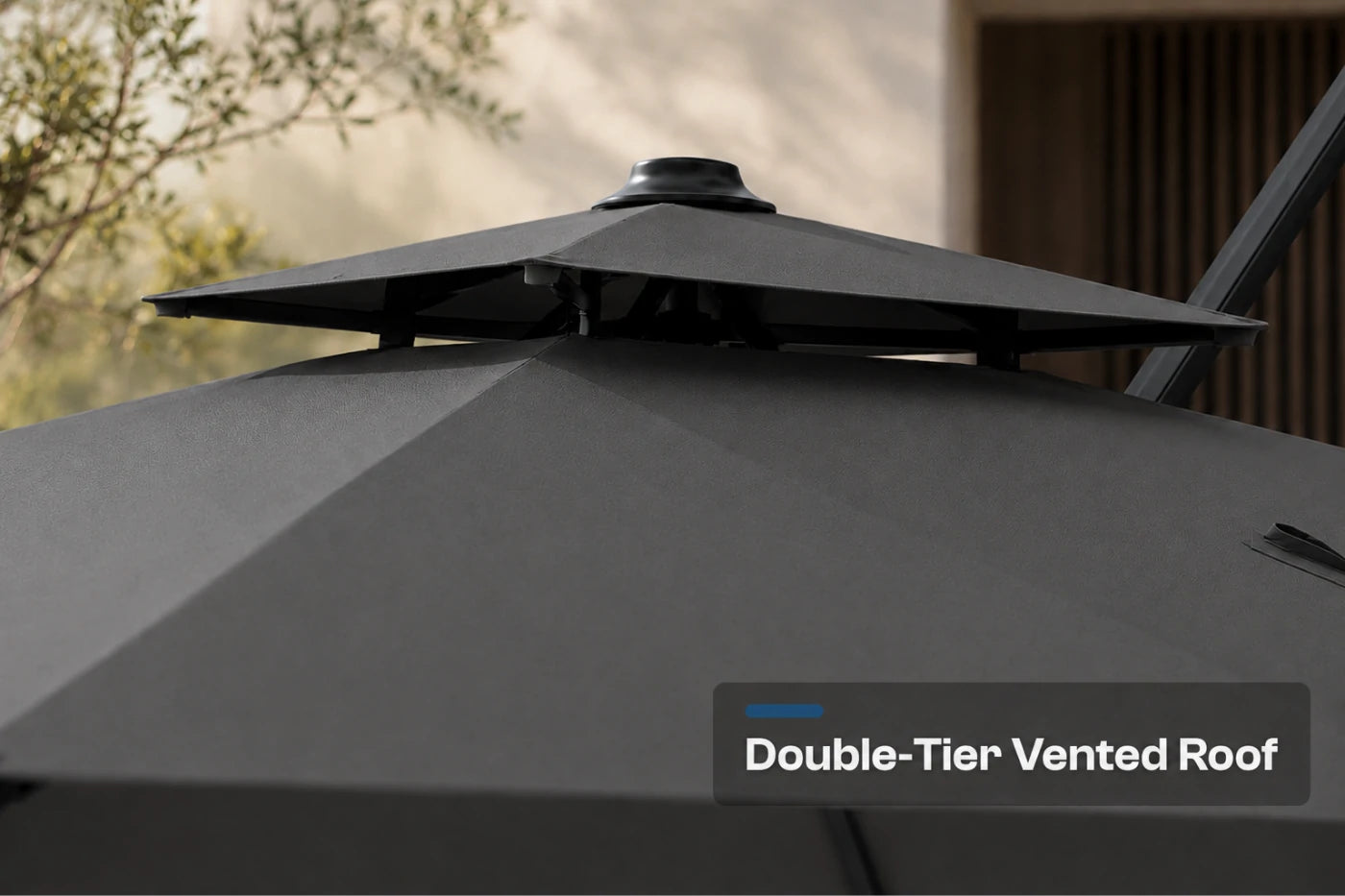 Close-up of a dark gray patio umbrella showing its double-tier vented roof structure, with a text overlay that reads 'Double-Tier Vented Roof' against an outdoor blurred background.