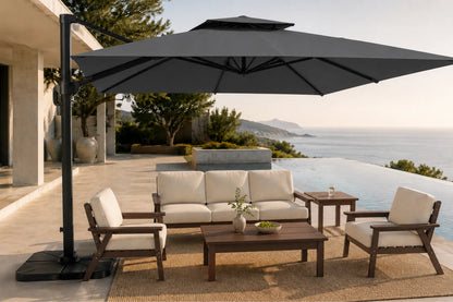 A large dark gray patio umbrella with a cantilevered pole stands over an outdoor seating area on a sunlit terrace overlooking a pool and sea. The furniture includes a cream cushioned sofa, two matching armchairs, a wooden coffee table, and a side table.