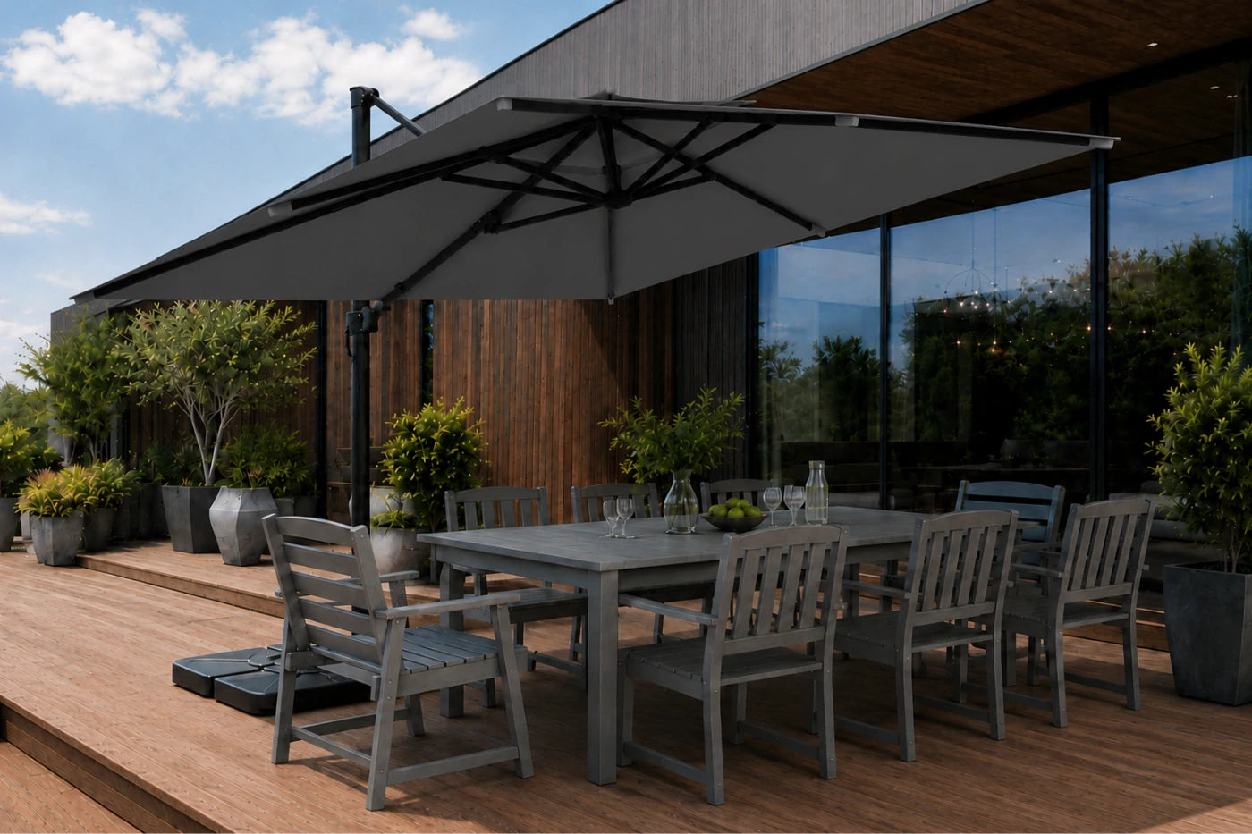 A large square patio umbrella in dark gray shades is shown on a wooden deck next to a modern outdoor dining table and chairs, with a house exterior and sky in the background.