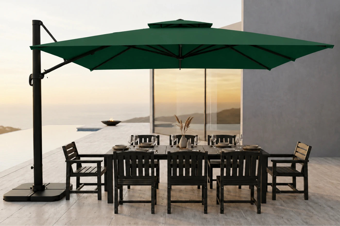A large dark green cantilever patio umbrella with a black base and pole, shading a dark outdoor dining table and chairs on a modern terrace overlooking a sunset seascape.