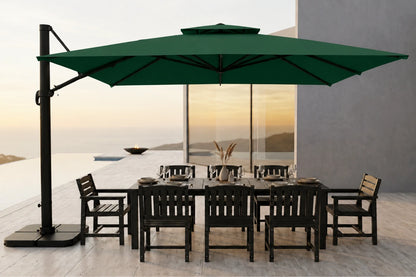 A large dark green cantilever patio umbrella with a black base and pole, shading a dark outdoor dining table and chairs on a modern terrace overlooking a sunset seascape.