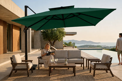 Outdoor patio furniture set with a large dark green cantilever umbrella, a three-seat sofa, two armchairs, and a coffee table on a poolside terrace. A woman sits on the sofa reading, and a man stands nearby, with a modern building and distant hills in the background.