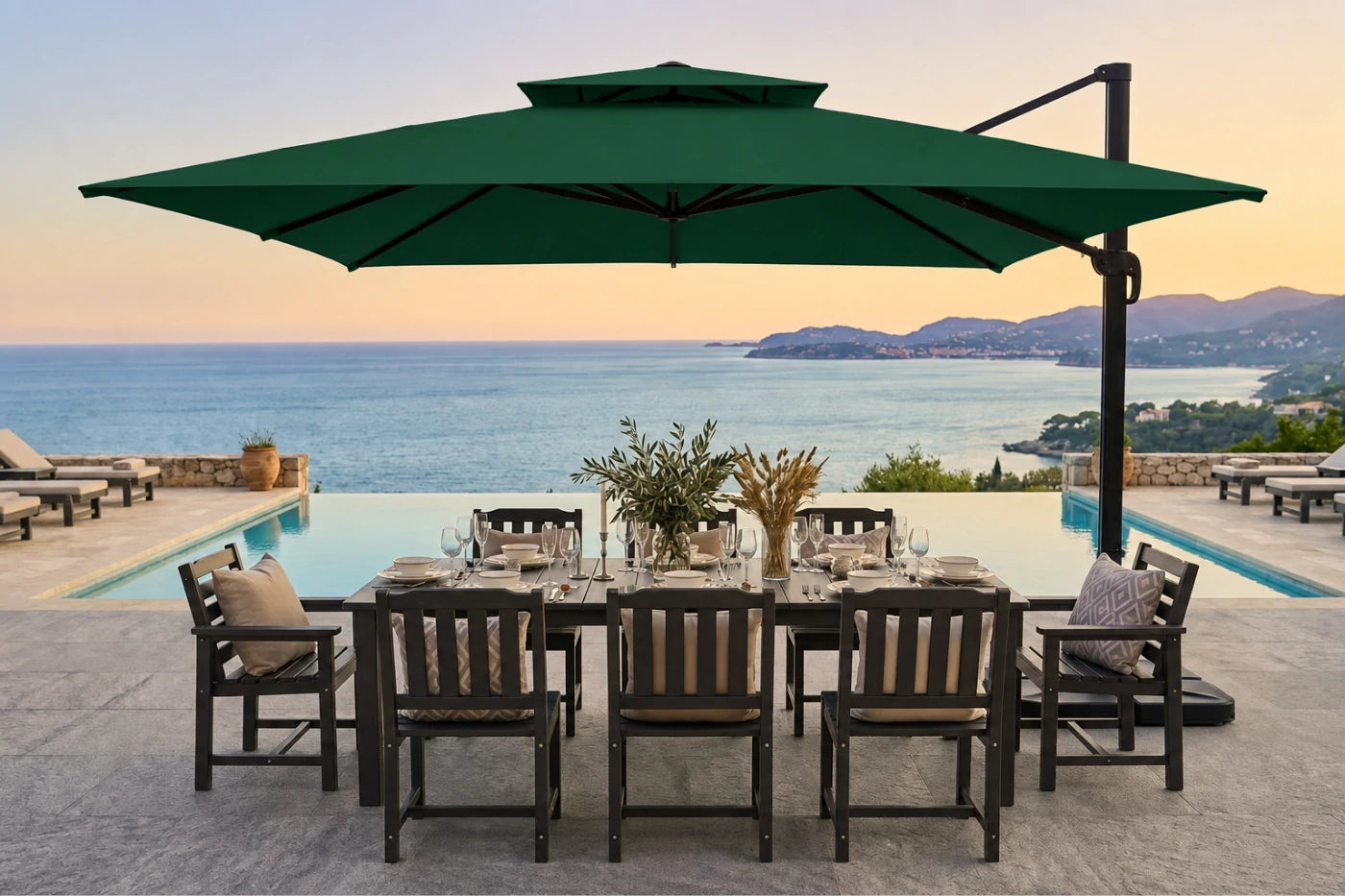 A large dark green patio umbrella with a cantilever arm over a dining set on a stone terrace overlooking an infinity pool and the sea at sunset.