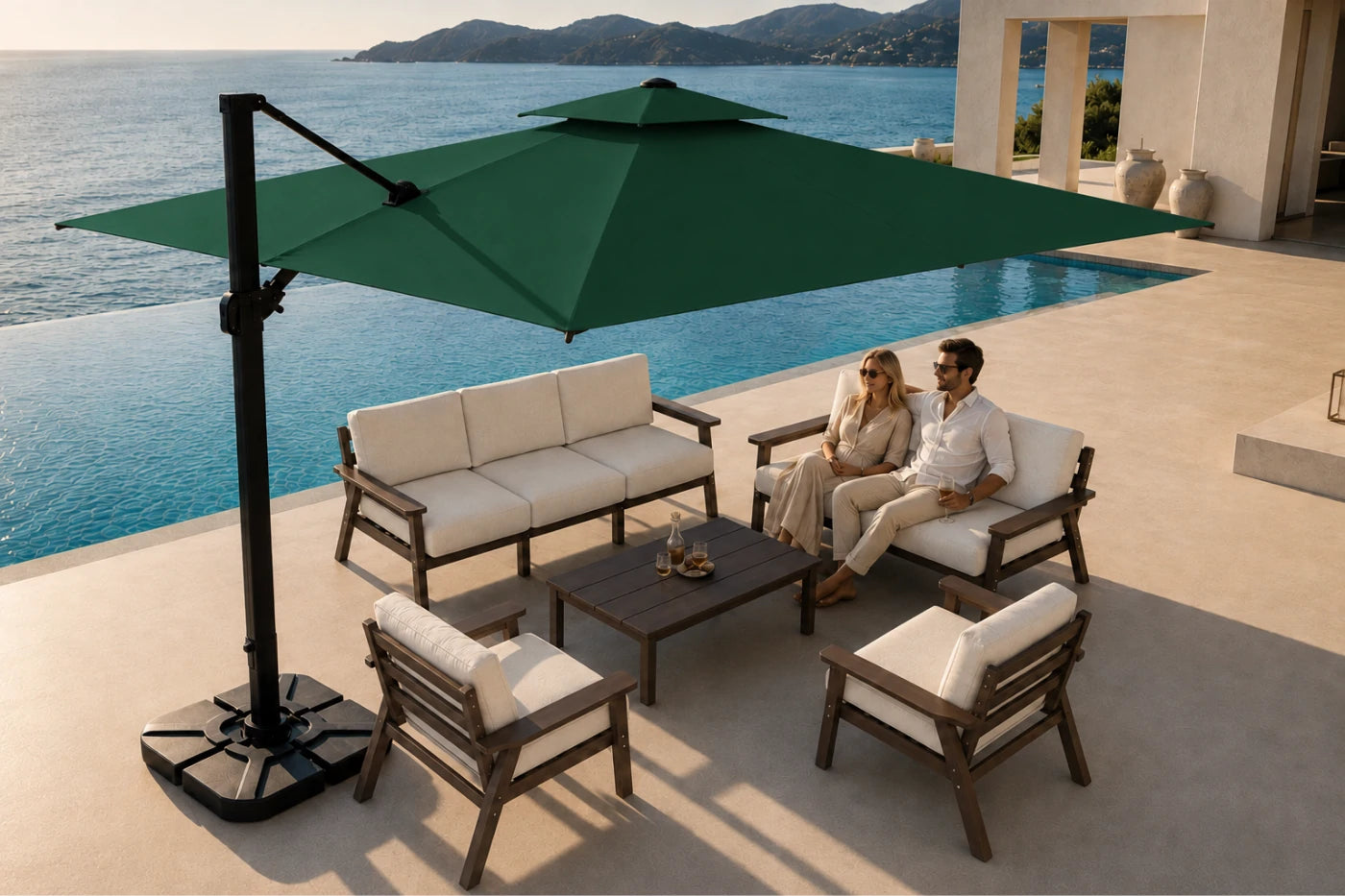Outdoor patio furniture set with a large dark green cantilever umbrella, a three-seat sofa, two armchairs, and a coffee table on a poolside terrace, with a couple sitting on the sofa. The background shows a swimming pool, ocean, and building architecture.