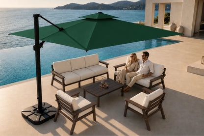 Outdoor patio furniture set with a large dark green cantilever umbrella, a three-seat sofa, two armchairs, and a coffee table on a poolside terrace, with a couple sitting on the sofa. The background shows a swimming pool, ocean, and building architecture.
