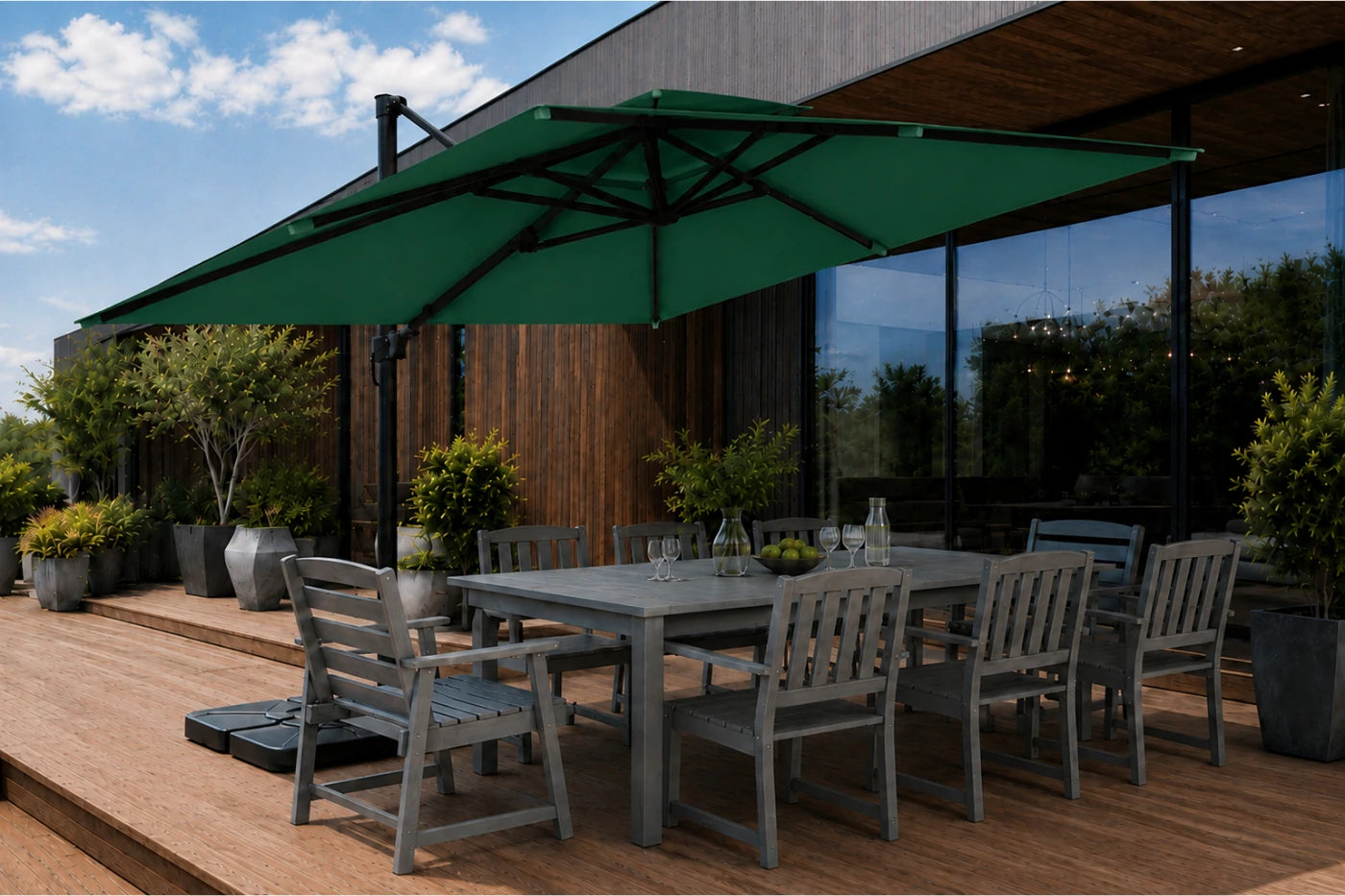 A dark green patio umbrella shading a gray outdoor dining table and chairs on a wooden deck beside a modern building with large windows.