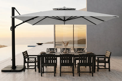 A large cantilever patio umbrella in gray with a black pole and base, shading a dark outdoor dining table and chairs on a modern terrace overlooking a sunset seascape.