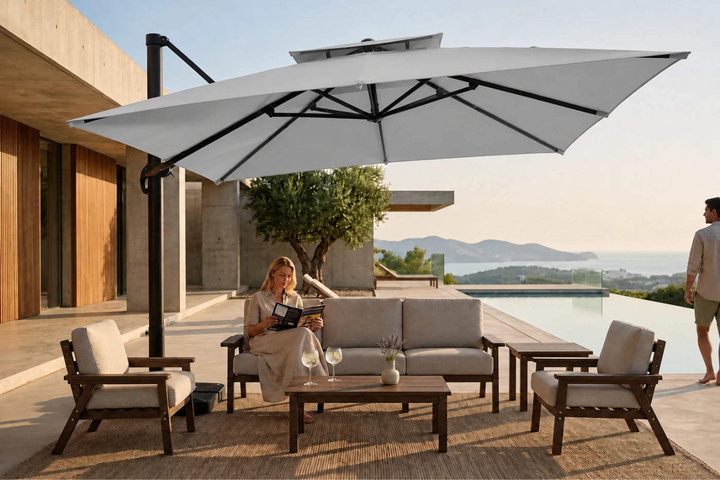 A large gray cantilever patio umbrella with a black pole is set up beside a modern outdoor lounge set on a poolside terrace. A woman sits on the sofa under the umbrella reading a magazine, and a man stands nearby.