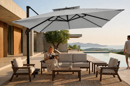 A large gray cantilever patio umbrella with a black pole is set up beside a modern outdoor lounge set on a poolside terrace. A woman sits on the sofa under the umbrella reading a magazine, and a man stands nearby.