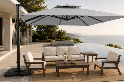 A luxury outdoor patio furniture set with a large gray cantilever umbrella, a three-seat sofa, two armchairs, and a coffee table on a woven rug, set against an infinity pool and ocean background.