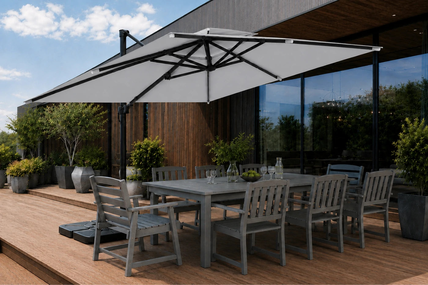 A large rectangular patio umbrella with a gray canopy and black frame, shading a gray outdoor dining table and chairs on a wooden deck beside a modern house with large glass windows.