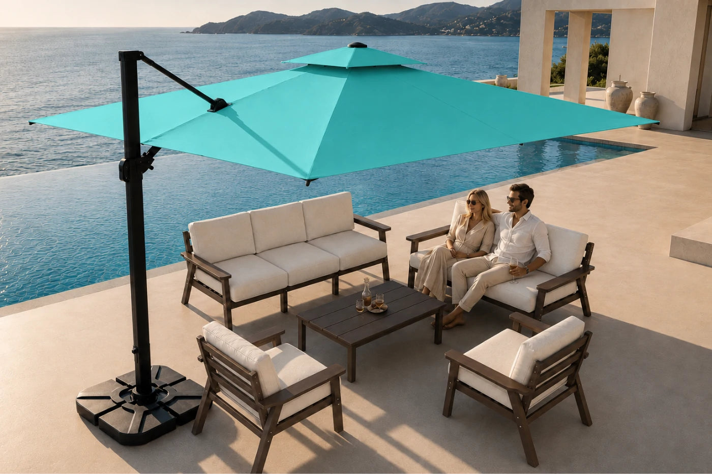 Outdoor patio furniture set with a large turquoise cantilever umbrella, a three-seat sofa, two armchairs, and a low coffee table on a sunlit terrace overlooking a pool and ocean. A man and woman are seated on the furniture.