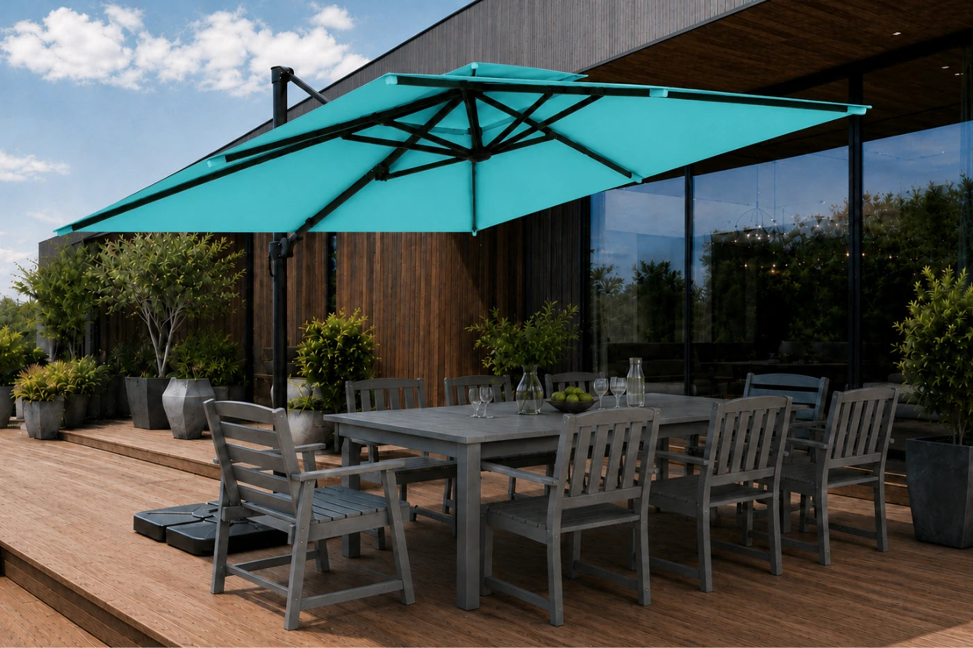 A large turquoise patio umbrella with a black frame shading a gray outdoor dining table and chairs on a wooden deck in front of a modern house.