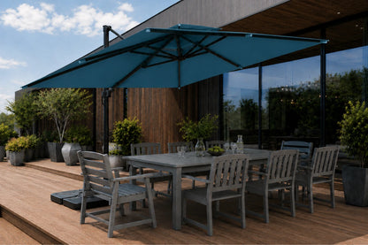 A large lake blue patio umbrella shading a gray outdoor dining table and chairs on a wooden deck beside a modern house with large windows.