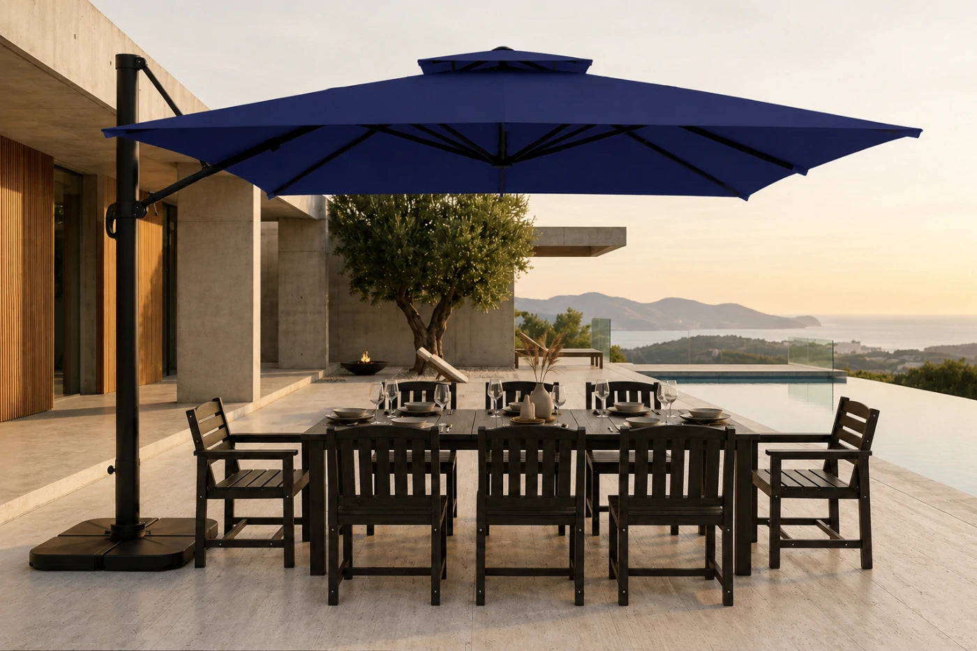 A large navy blue cantilever patio umbrella with a black metal frame and base, shading a dark outdoor dining table and chairs on a modern terrace overlooking a pool and distant mountains.