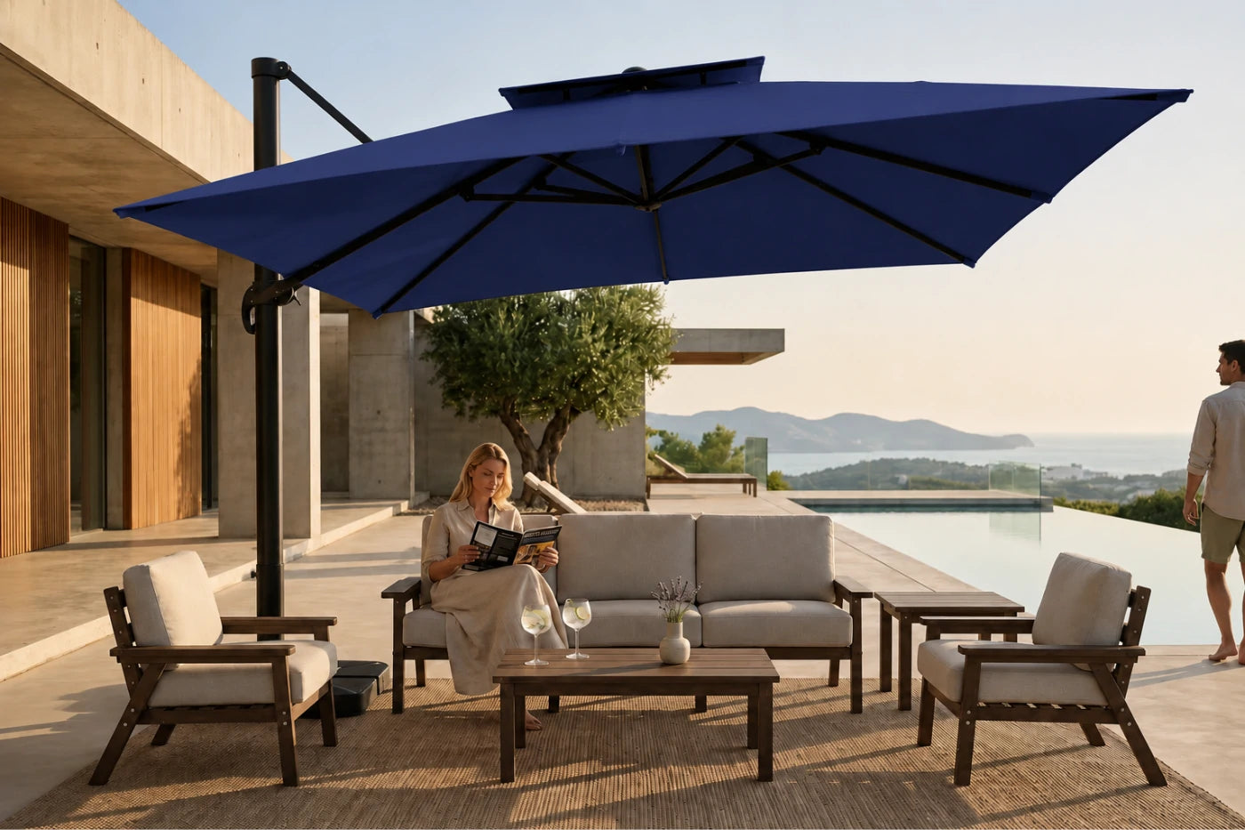 A large navy blue cantilever patio umbrella in a modern outdoor patio setting, shading a three-seater sofa, two armchairs, and a coffee table. A woman is seated on the sofa, and a man is standing on the right edge of the patio overlooking a pool and mountains.