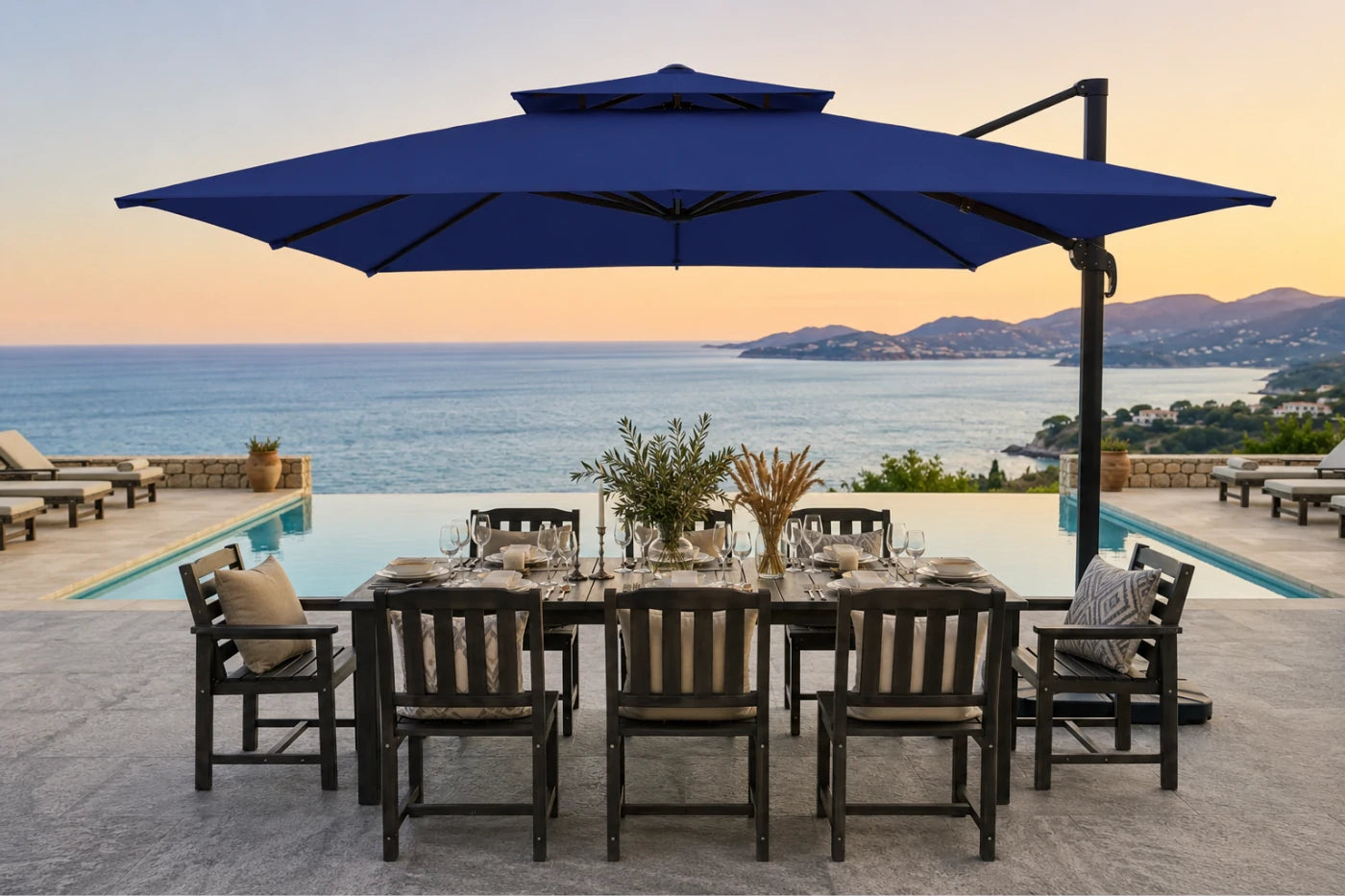A large navy blue cantilever patio umbrella over a dining table and chairs, set on a stone patio with an infinity pool and ocean view at sunset.