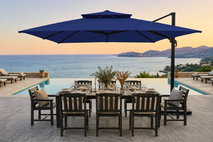 A large navy blue cantilever patio umbrella over a dining table and chairs, set on a stone patio with an infinity pool and ocean view at sunset.