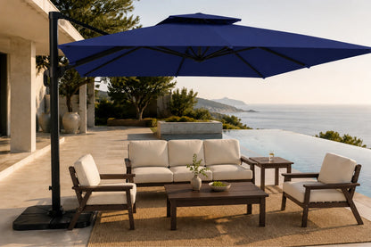 A large navy blue patio umbrella with a black cantilever pole stands over an outdoor living set on a poolside terrace. The set includes a cream cushioned sofa, two matching armchairs, a dark wood coffee table, and a side table, with a modern house and ocean in the background.