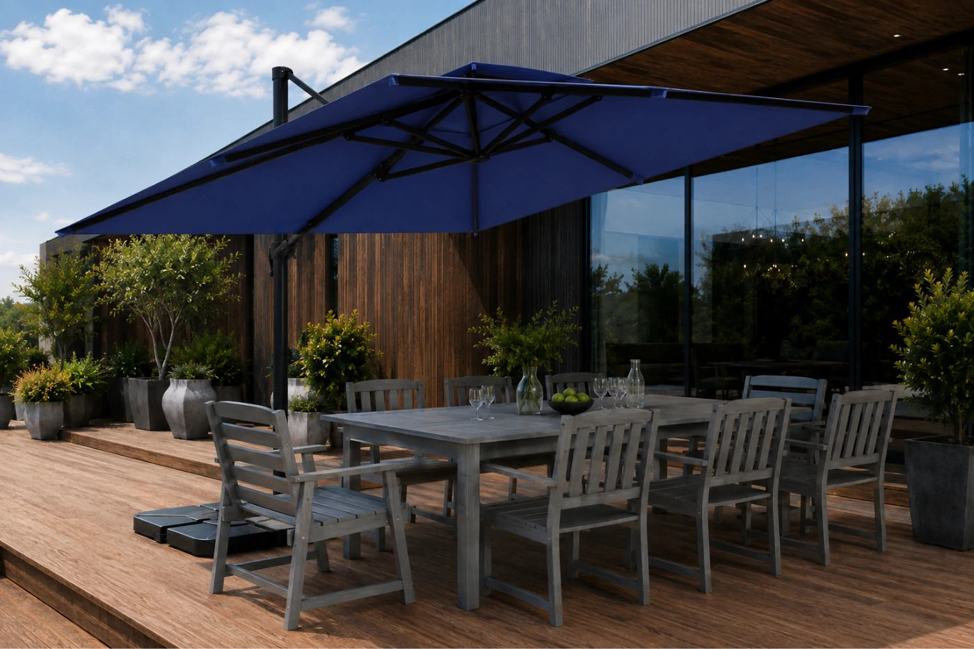 A large navy blue patio umbrella with a black frame shading a gray outdoor dining table and chairs on a wooden deck next to a modern glass-walled building.