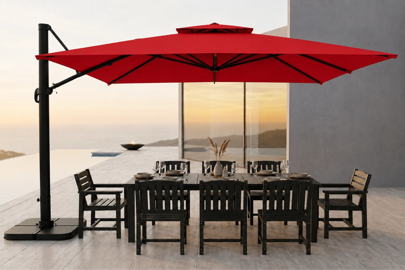 A large red cantilever patio umbrella with a black pole and base, shading a dark outdoor dining table and chairs on a modern terrace overlooking a sunset seascape.
