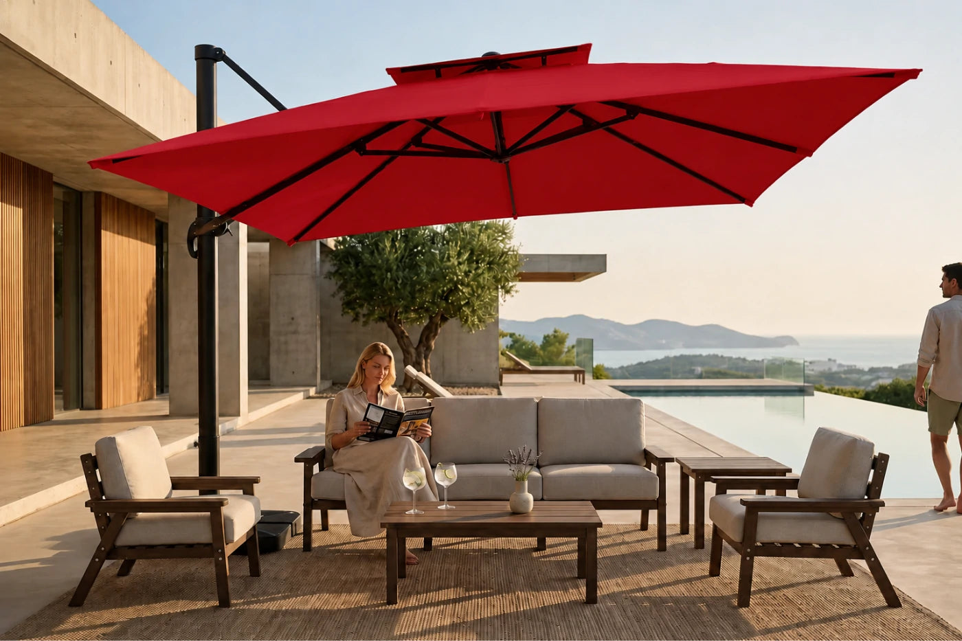A large red cantilever patio umbrella shades a modern outdoor seating area with a sofa, two armchairs, and a coffee table on a poolside terrace. A woman sits on the sofa reading, and a man stands nearby.