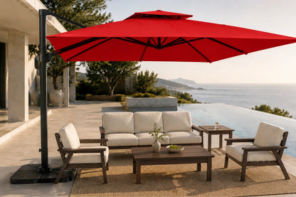 A luxury outdoor patio setup featuring a large red cantilever patio umbrella, a three-seater sofa, two armchairs, a coffee table, and a side table on a woven rug, set against an infinity pool and ocean backdrop.