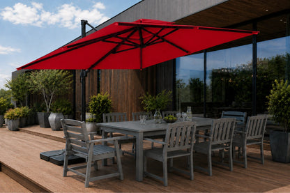 A large red patio umbrella with a black frame shading a gray outdoor dining table and chairs on a wooden deck beside a modern glass-walled building.