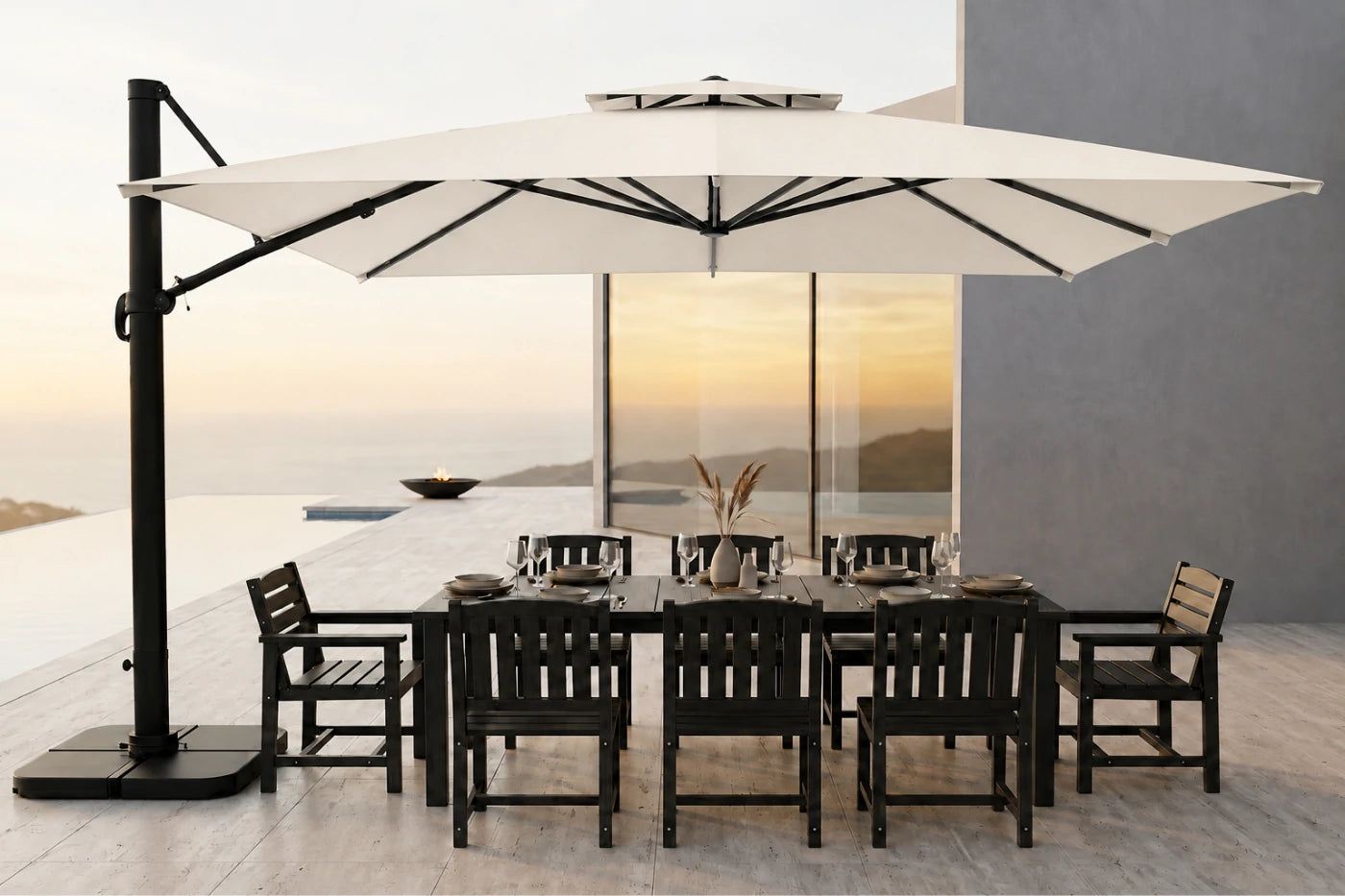 A large cantilever patio umbrella in creamy white with a black metal frame and base, shading a dark outdoor dining table and chairs on a modern terrace overlooking a sunset over water.