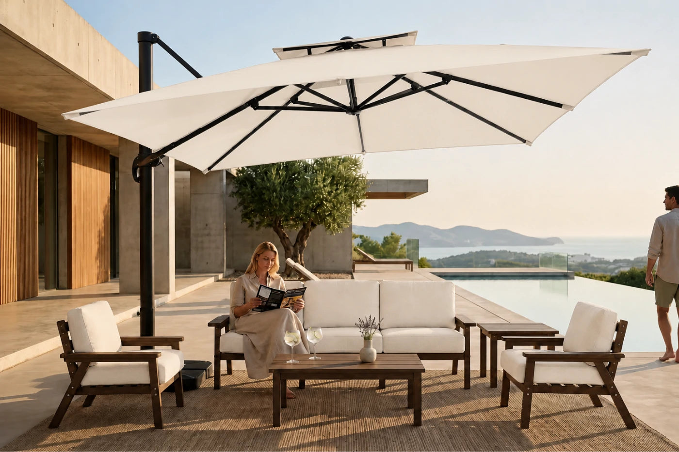 A large creamy white double canopy patio umbrella with a black offset pole is shown in a luxurious outdoor patio setting. Under the umbrella, a woman sits on a white cushioned three-seater sofa with a wooden coffee table.