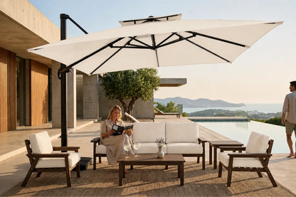 A large creamy white double canopy patio umbrella with a black offset pole is shown in a luxurious outdoor patio setting. Under the umbrella, a woman sits on a white cushioned three-seater sofa with a wooden coffee table.