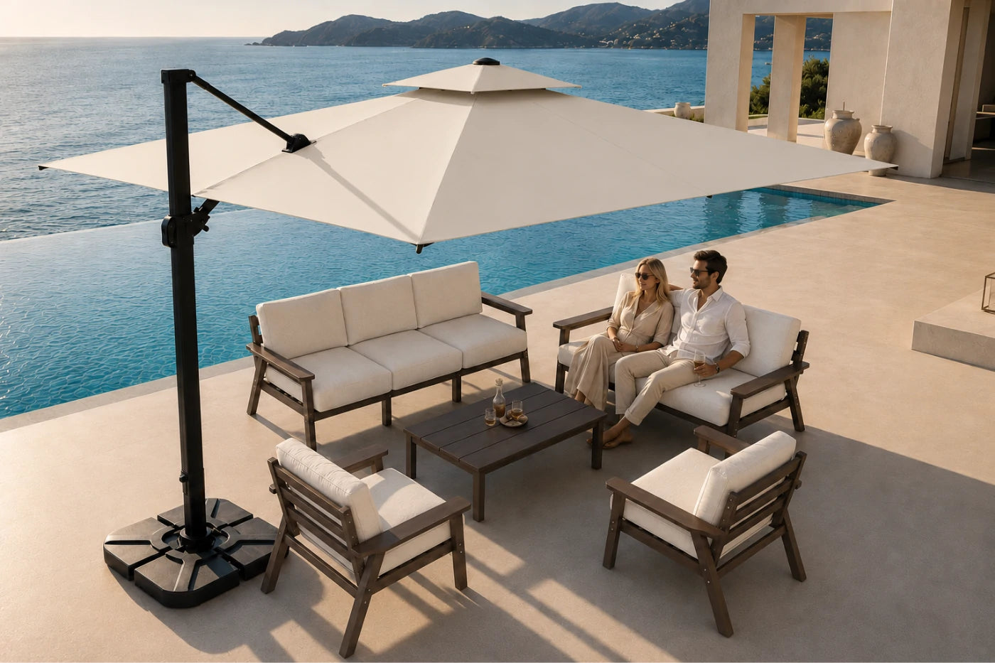 Outdoor patio furniture set with a large cream cantilever umbrella, a three-seat sofa, two armchairs, and a low rectangular coffee table, arranged beside a pool with a couple sitting on the sofa. The furniture has dark brown wood frames and off-white cushions.