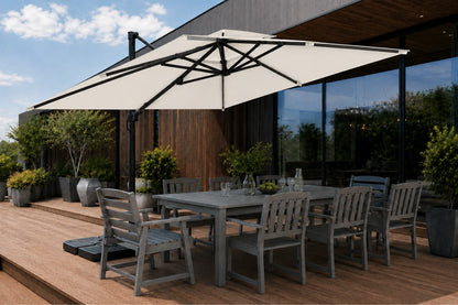 A large creamy white patio umbrella with a black frame and pole, shading a gray outdoor dining table and chairs on a wooden deck next to a modern house with large glass windows.