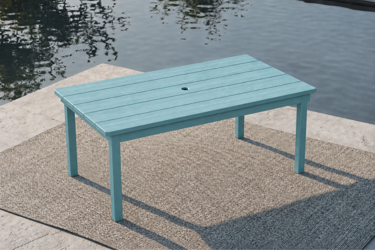 An Aruba blue rectangular outdoor dining table with a slatted top and a central hole, set on a textured patio rug next to a body of water.