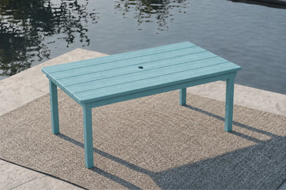 An Aruba blue rectangular outdoor dining table with a slatted top and a central hole, set on a textured patio rug next to a body of water.