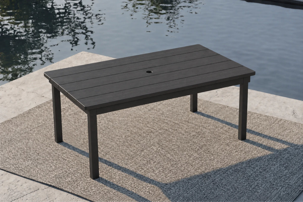A rectangular outdoor dining table with a black finish, featuring a central umbrella hole, set on a textured patio rug next to a body of water.