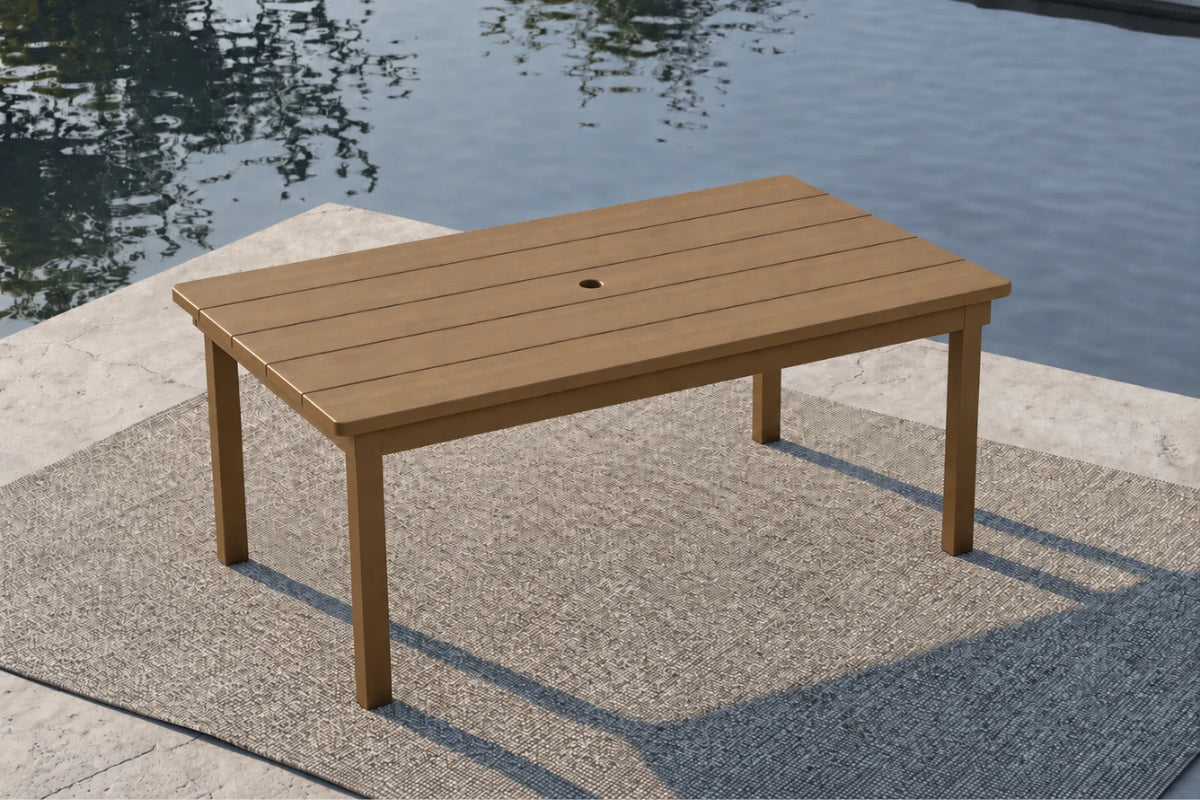 A rectangular outdoor table with a slatted wood-look top and four legs, positioned on a textured patio rug beside a body of water.