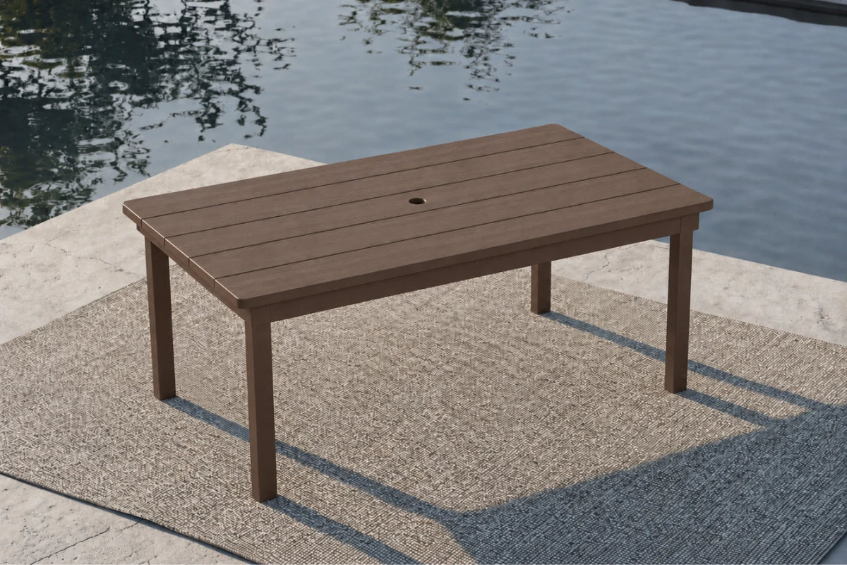 A rectangular outdoor table with a coffee-colored slatted top and matching legs, positioned on a textured patio surface next to a body of water.