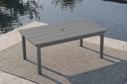 A rectangular outdoor table with a gray slatted top and matching legs, positioned on a textured patio rug beside a body of water.