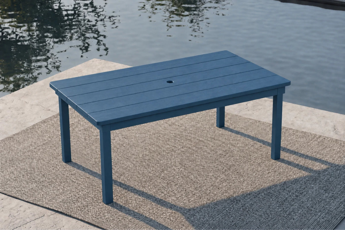 A rectangular blue outdoor table with a central hole, set on a textured patio rug next to a body of water.