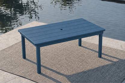 A rectangular blue outdoor table with a central hole, set on a textured patio rug next to a body of water.
