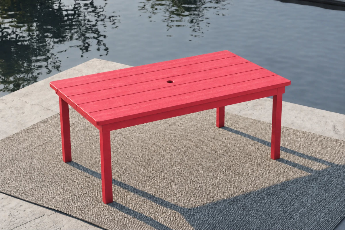 A red rectangular outdoor table with a slatted top and four legs, positioned on a textured gray rug beside a body of water.