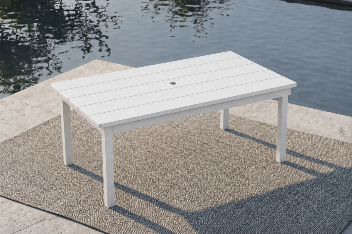 A white rectangular outdoor patio table with a slatted top and four legs, positioned on a textured rug beside a body of water.