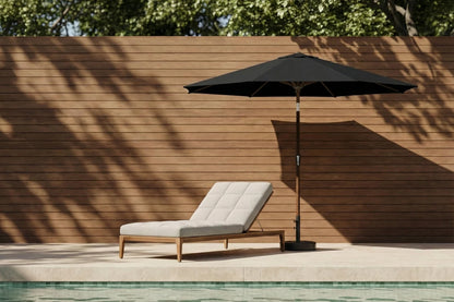 Chaise lounge with a black umbrella by a poolside against a wooden wall.