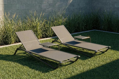 Two beige lounge chairs on grass with a stone wall and plants in the background